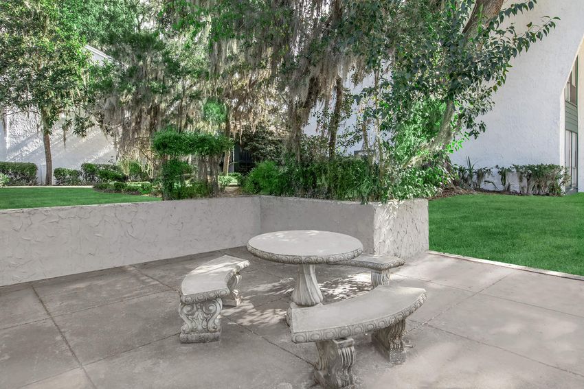 A serene outdoor setting featuring a concrete table with matching benches, surrounded by lush greenery and trees. The area is well-maintained, with soft grass and decorative plants, creating a peaceful atmosphere ideal for relaxation or social gatherings.