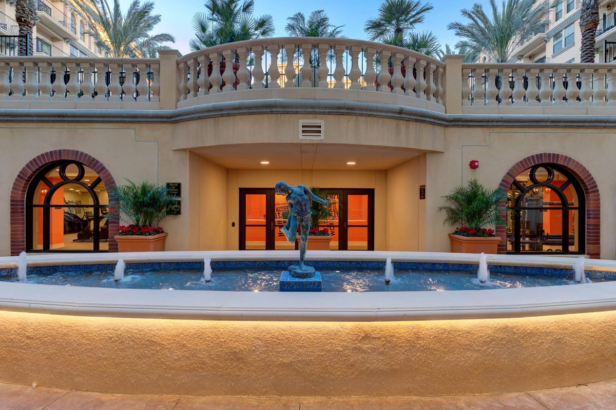 A decorative fountain featuring a blue sculpture of a standing figure, surrounded by potted plants. The background includes elegant arches and windows of a building, under palm trees, creating a serene and upscale atmosphere. Soft lighting enhances the ambiance.