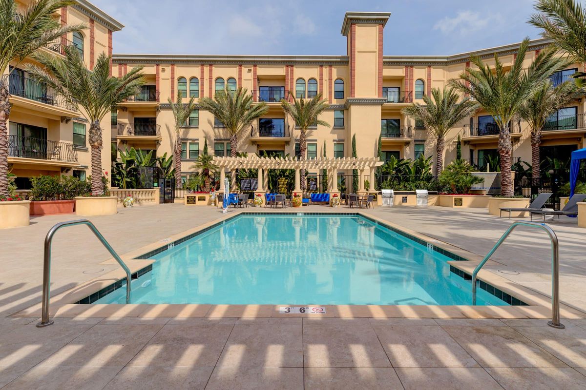 A beautifully designed outdoor pool area surrounded by palm trees, lounge chairs, and greenery. The pool has a clear blue water and is situated in front of a multi-story building with balconies. The setting exudes a relaxed, tropical vibe under a partly cloudy sky.