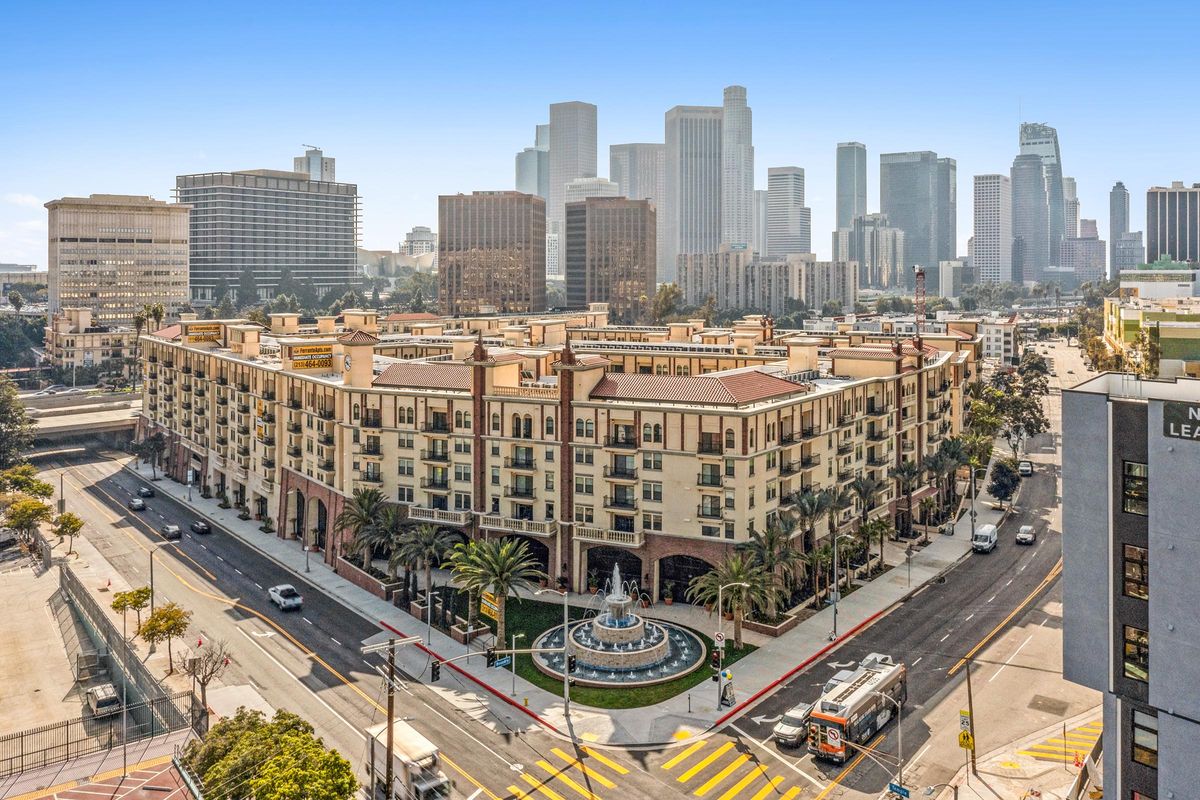 Aerial view of a cityscape featuring a large, ornate building with a fountain in the center of a circular plaza. Surrounding the plaza are palm trees and bustling streets. In the background, modern skyscrapers of a downtown area rise against a clear blue sky.