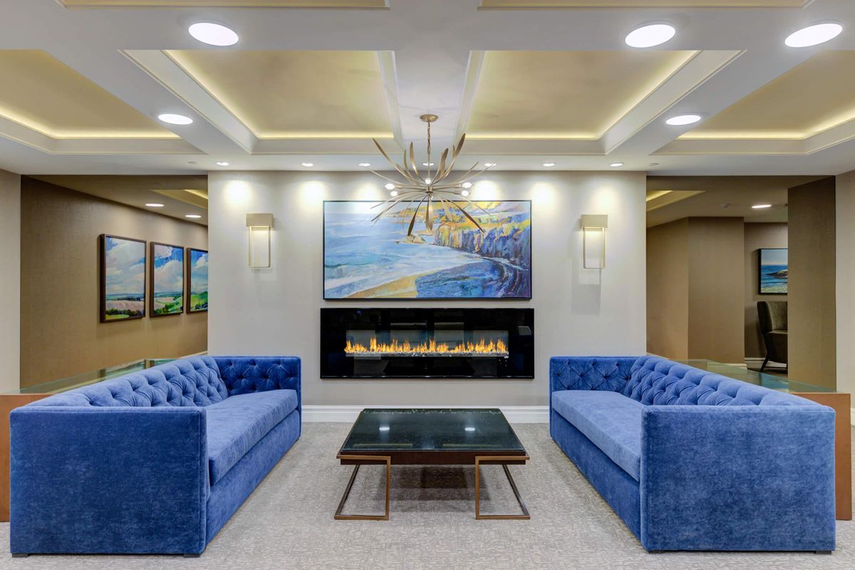 A modern lounge featuring two blue sofas facing each other across a black glass coffee table. Above, a contemporary chandelier hangs from the ceiling, and a large painting of a coastal landscape is displayed on the wall. A sleek electric fireplace adds warmth to the space, which is enhanced by recessed lighting.