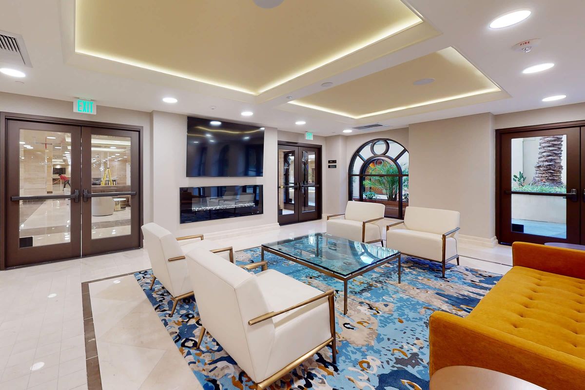 A modern lobby area featuring a glass coffee table on a vibrant rug, surrounded by white armchairs and an orange couch. Large windows and double doors lead outside, with a contemporary wall-mounted TV and a fireplace adding to the ambiance. Soft lighting enhances the inviting atmosphere.