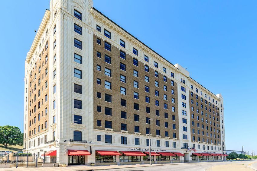 A large, multi-story brick building with a white facade. The structure features numerous windows and a prominent entrance with red awnings. The sky is clear blue, and the building is situated on a wide street, indicating an urban setting. Trees are visible in the background.
