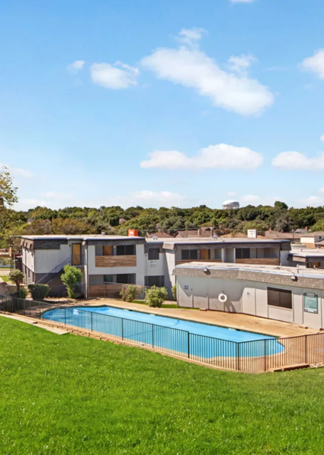 Spacious apartment complex with a refreshing outdoor pool, set against a backdrop of lush greenery and clear blue skies.