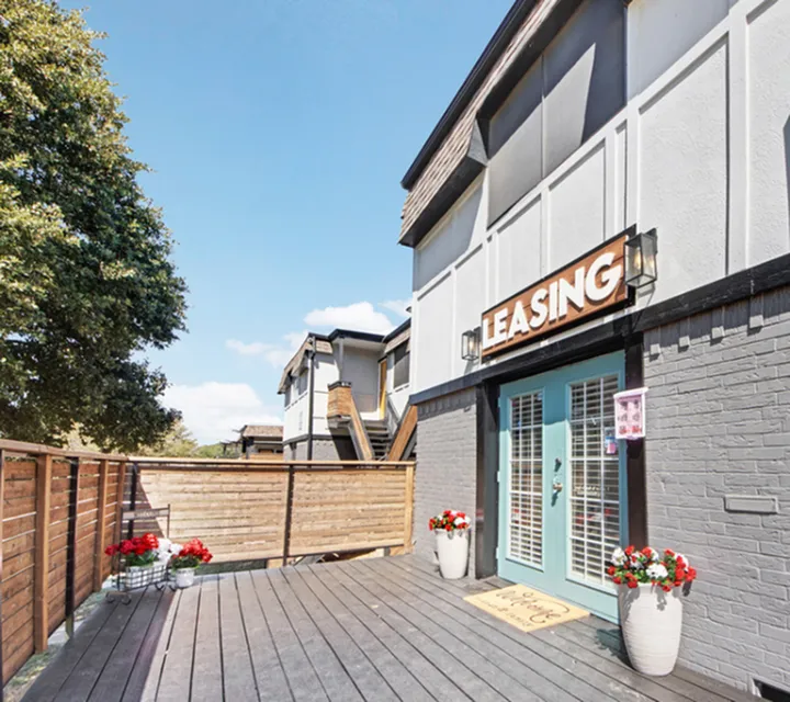 Charming apartment complex with modern leasing office, bright entrance, and inviting outdoor deck, enhanced by vibrant potted flowers.