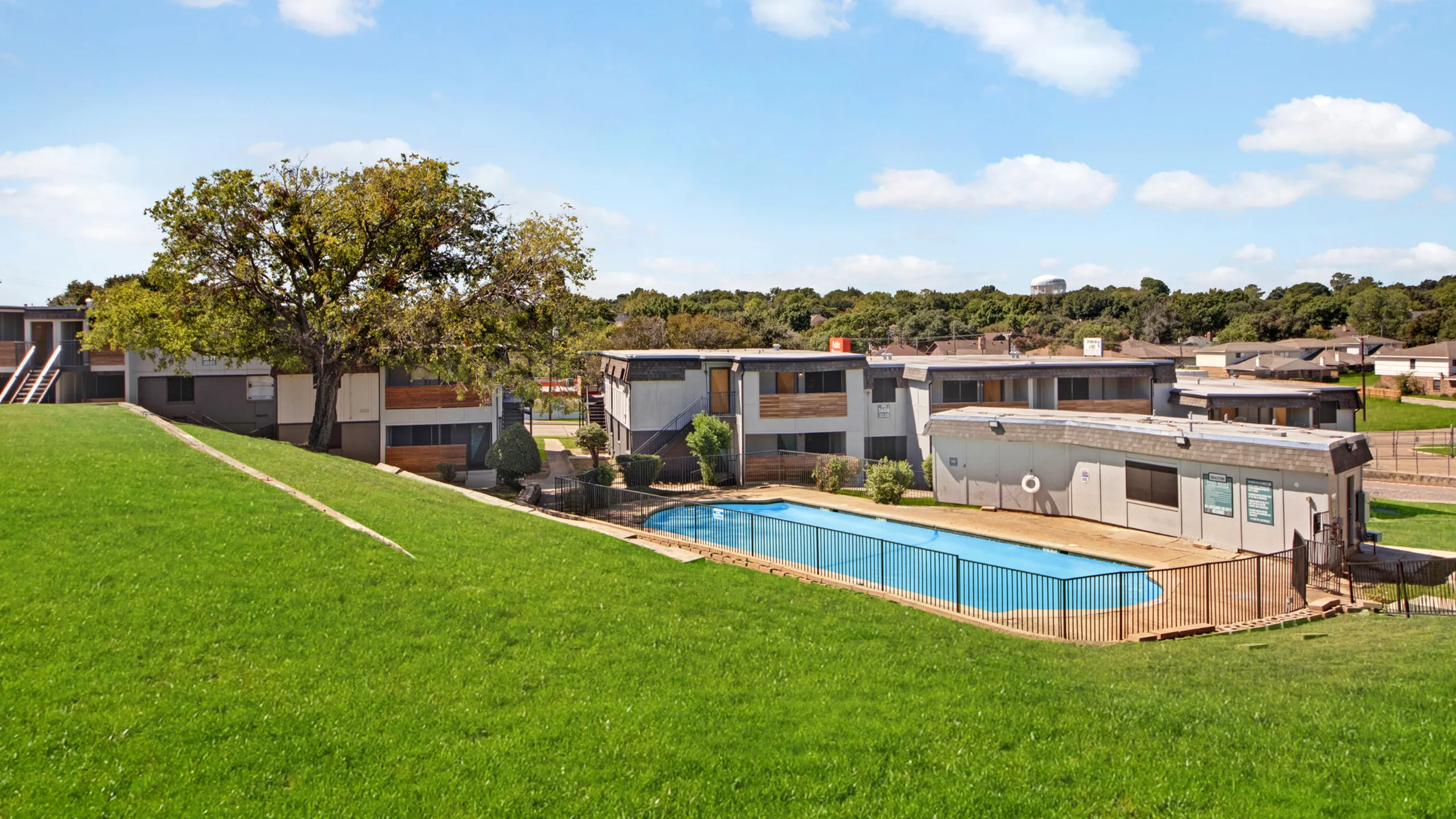 Inviting apartment complex with a large swimming pool, surrounded by lush green lawns and modern architecture under a bright blue sky.