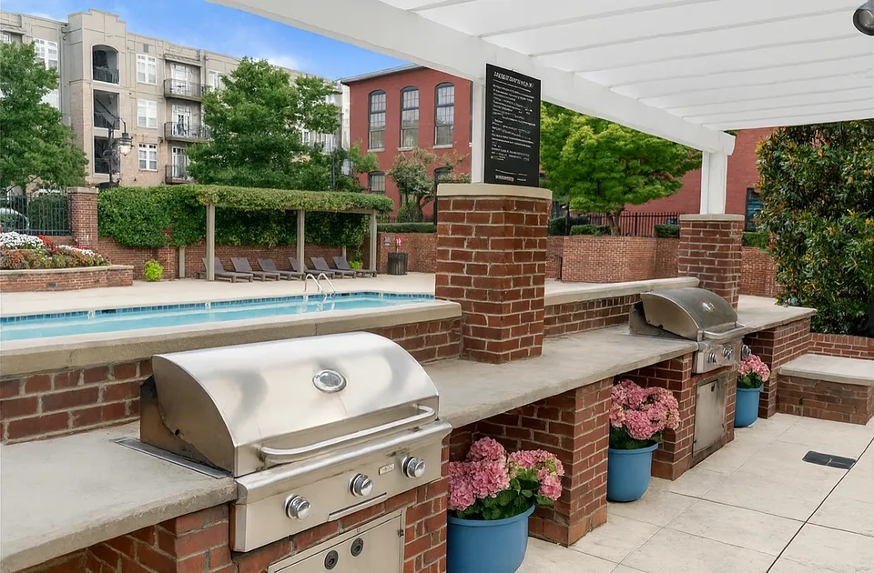 Charming outdoor space featuring sleek stainless steel grills, vibrant flower planters, and a refreshing swimming pool with stylish lounging chairs, surrounded by lush greenery and elegant brick architecture at Alpha Mill Apartments in Charlotte NC