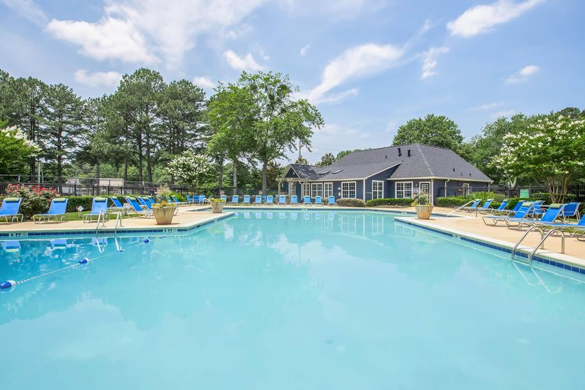 A large outdoor swimming pool surrounded by lounge chairs and potted plants. Lush green trees line the background, and a blue sky with scattered clouds completes the serene atmosphere. A clubhouse is visible nearby, creating a relaxing recreational space.