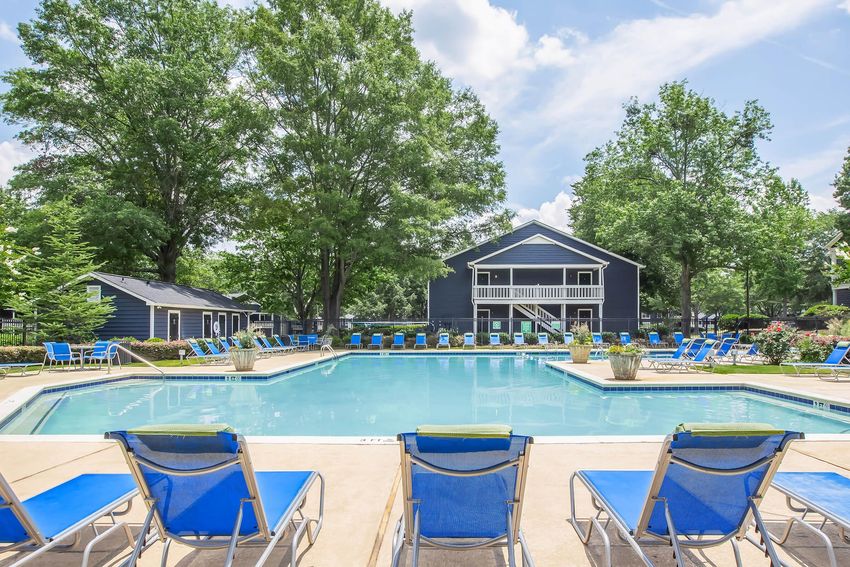 A clear blue swimming pool surrounded by lounge chairs, with lush green trees in the background. A two-story building with a balcony overlooks the pool area, and the sky is bright with fluffy white clouds. The scene conveys a relaxed and inviting outdoor atmosphere.