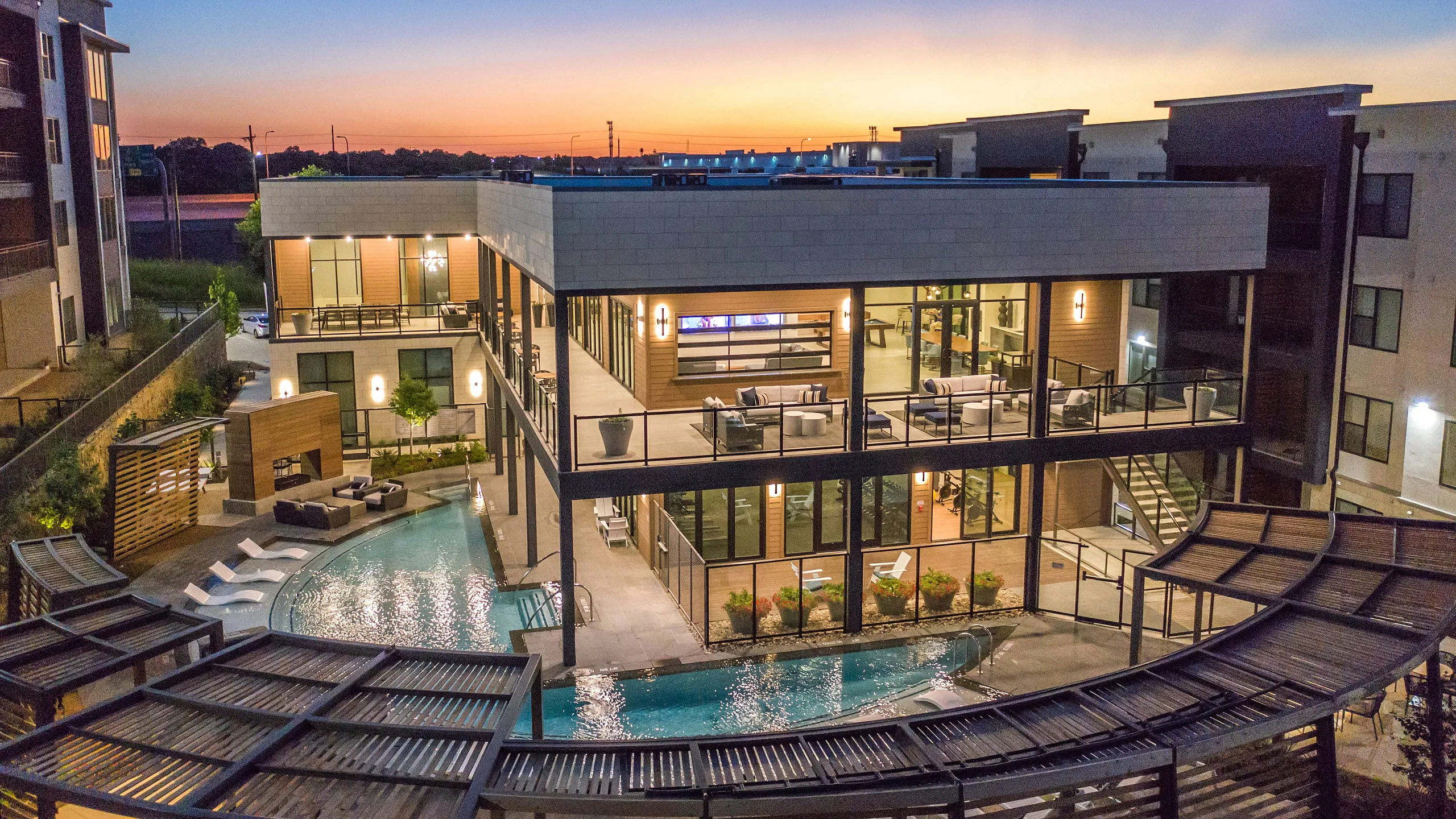 aerial view of the two story clubhouse brightly lit at twilight over the crescent shaped pool