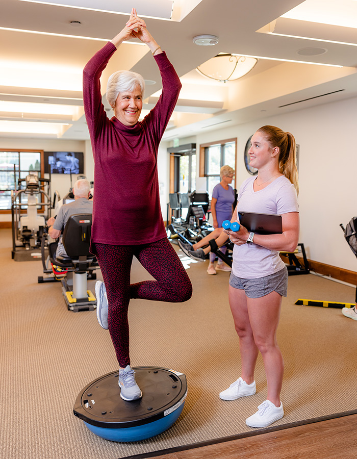 A senior woman on a balancing ball in a gym with a trainer.