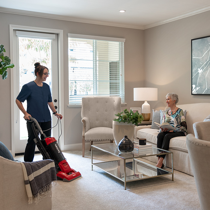 A housekeeper vacuuming while an elderly woman sits on the sofa reading.