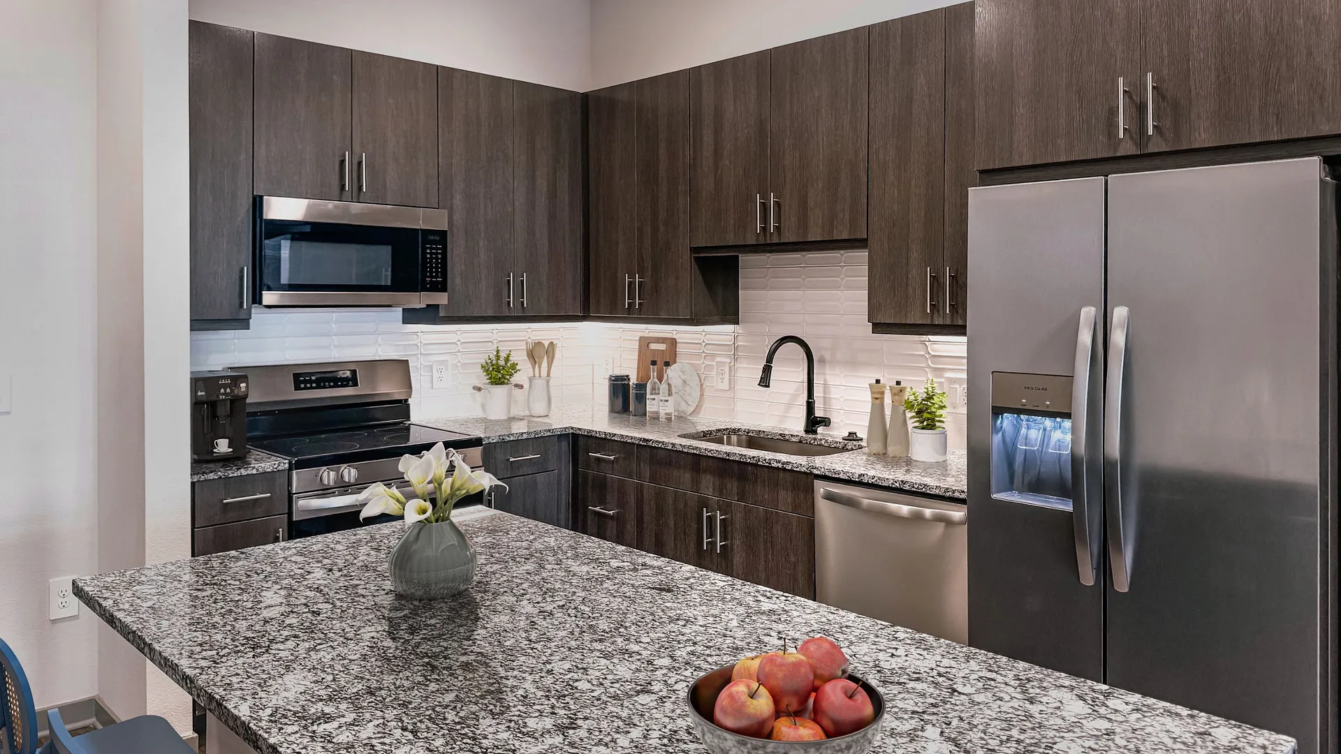 Modern kitchen with dark cabinets, stainless steel appliances, and a granite countertop featuring a bowl of apples.