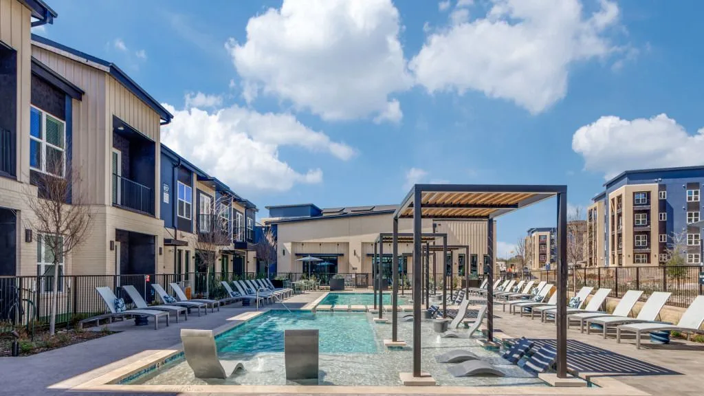 A modern apartment complex featuring a spacious pool area with lounge chairs and a sunny sky.