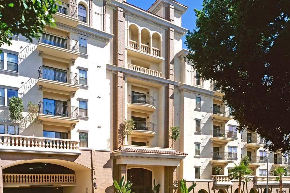 A modern multi-story apartment building featuring balconies, arched architectural elements, and lush greenery surrounding the entrance. The sky is clear and blue.