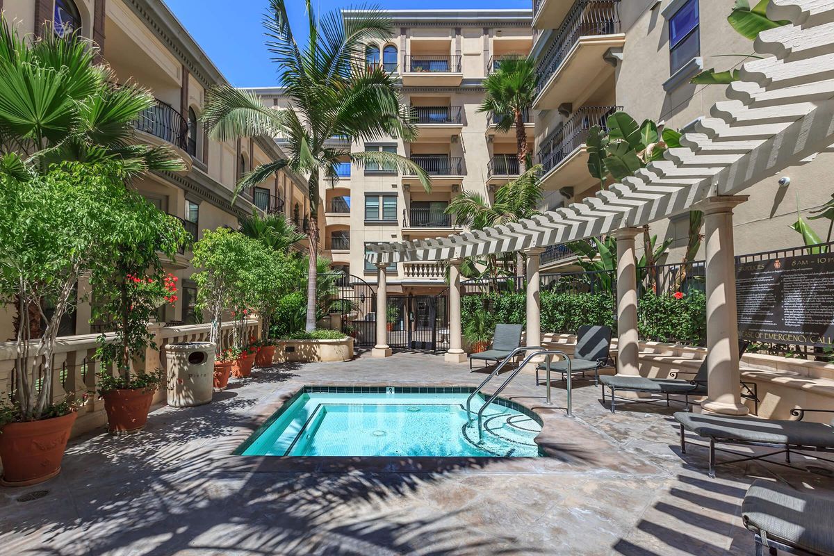 A well-maintained courtyard with a small swimming pool surrounded by lounge chairs and greenery, featuring palm trees and potted plants, under a bright blue sky.