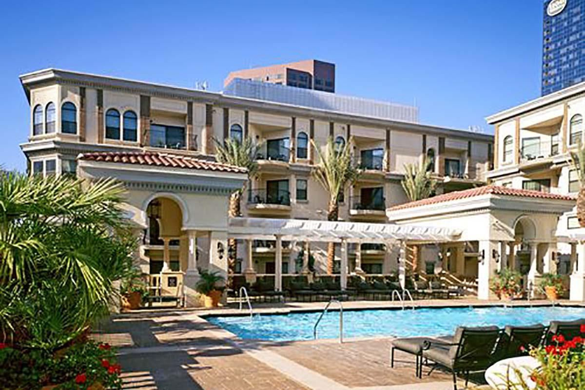 A spacious outdoor pool area surrounded by palm trees and lounge chairs, with a multi-story building in the background under a clear blue sky.