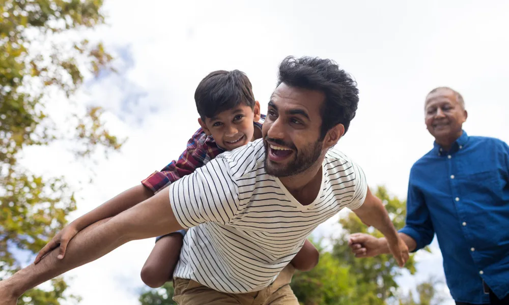 smiling man with child on back with older man in background outdoors in daytime