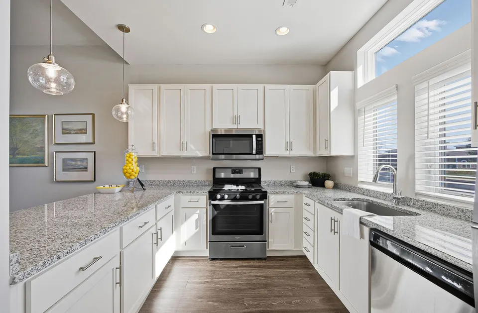 Bright and modern kitchen with sleek white cabinets, gleaming granite countertops, and stainless steel appliances, highlighted by elegant pendant lighting and large windows.
