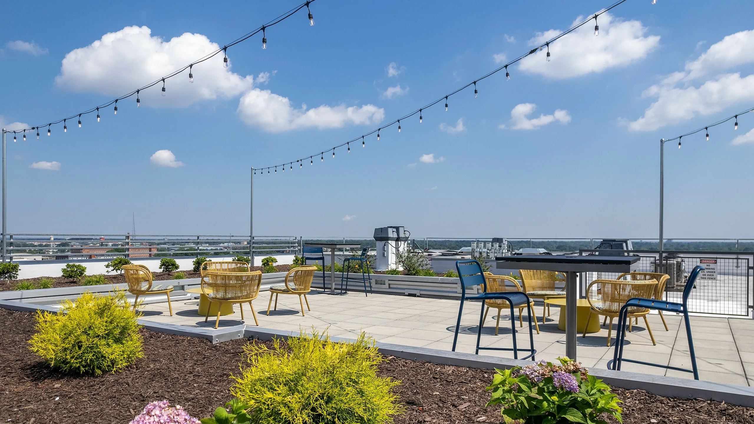 Modern rooftop terrace with stylish seating, string lights, and vibrant landscaping under a clear blue sky.