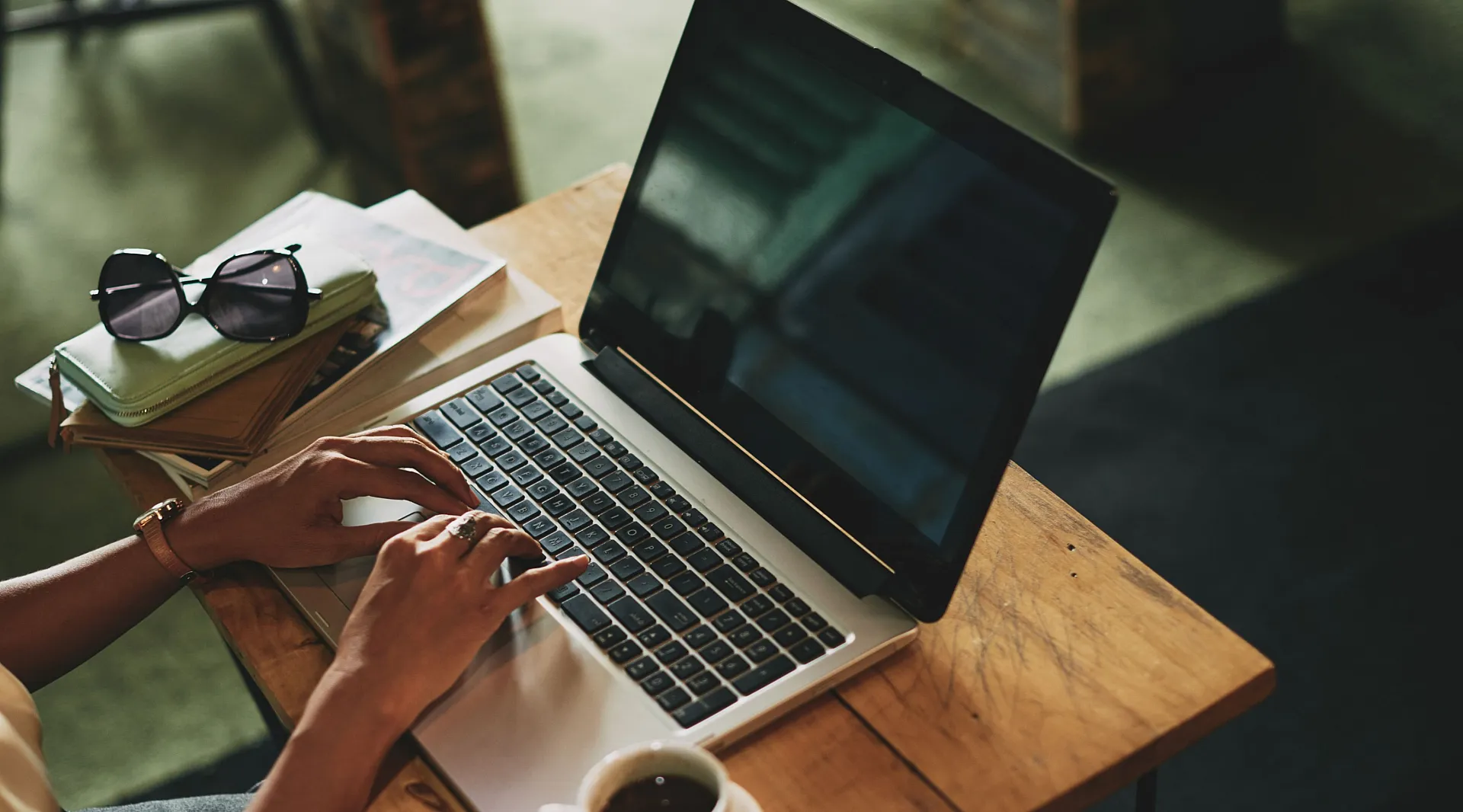 Cozy workspace with someone typing on a laptop at a wooden table, accompanied by a stack of books, sunglasses, and a coffee cup.
