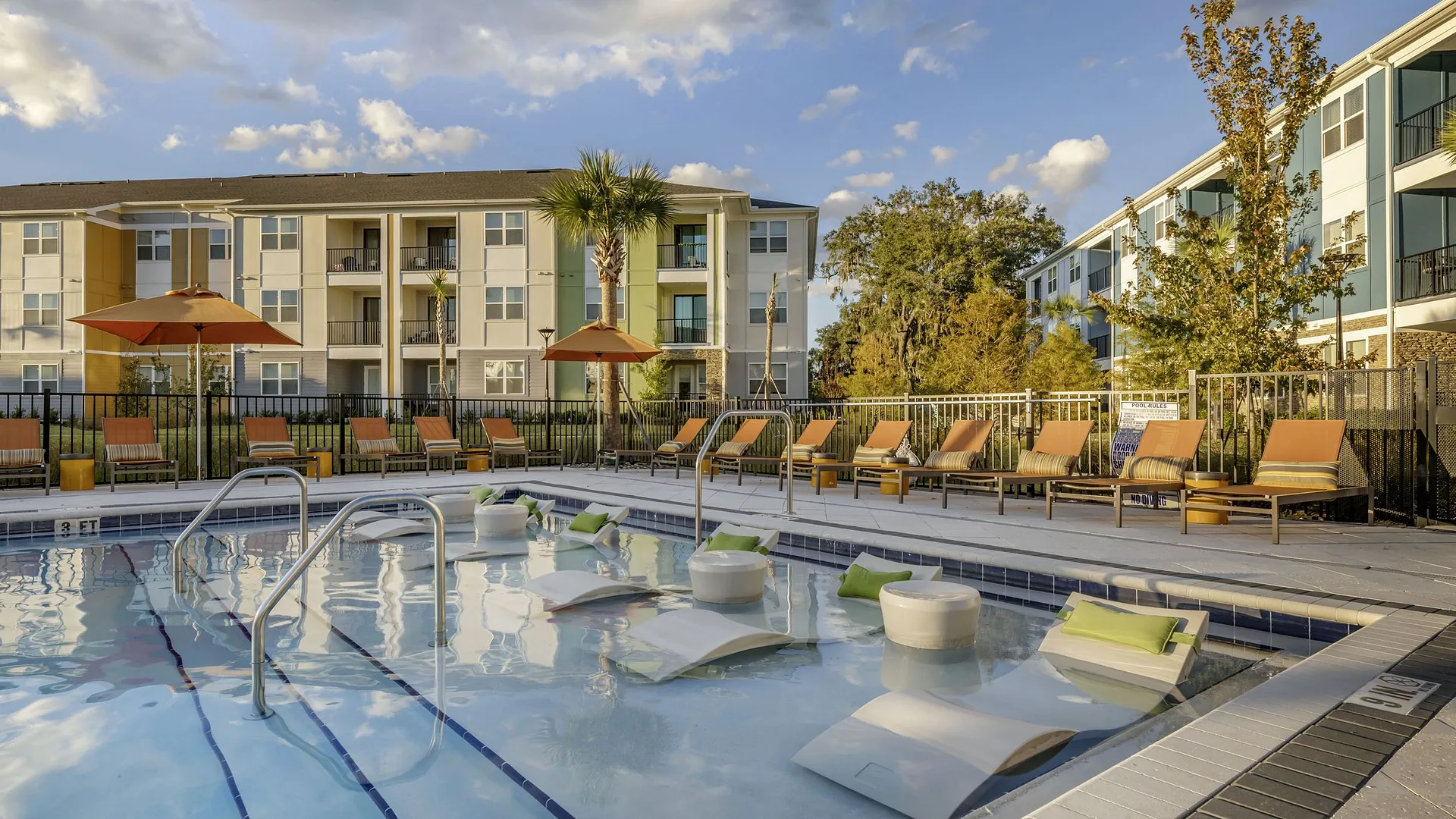 A sunny pool area with lounge chairs and umbrellas, surrounded by modern apartment buildings.