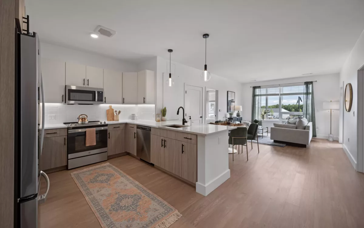 Open kitchen with stainless steel appliances, wood cabinets, black plumbing and fixtures, and bar seating, leading to a living area.