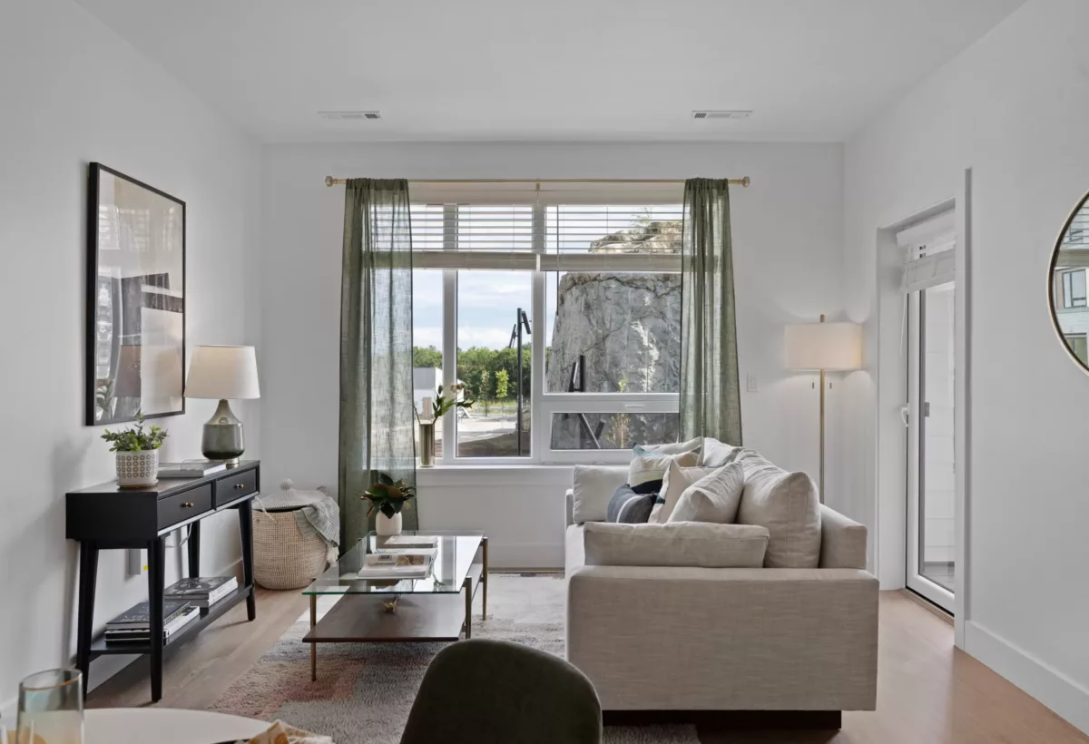 Bright living room with a grey sofa, large window, glass coffee table with books, and a console table with decor.