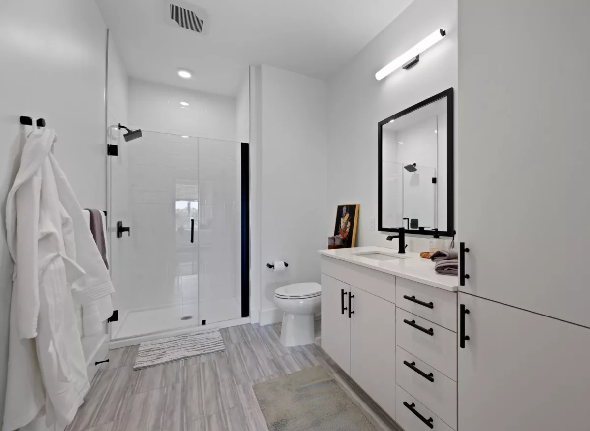 Bright bathroom with glass shower, white cabinets, large mirror, and wood-style flooring.