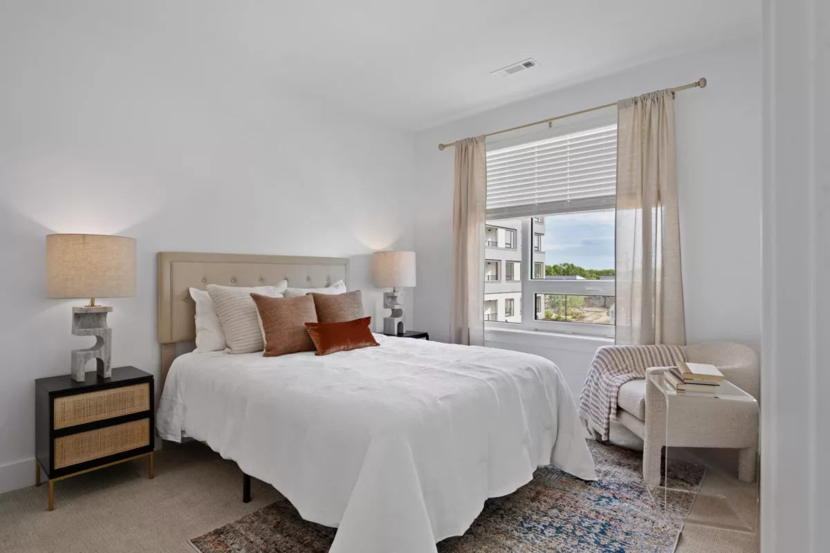 Bedroom with beige and brown pillows, two lamps, and a window overlooking an apartment complex.