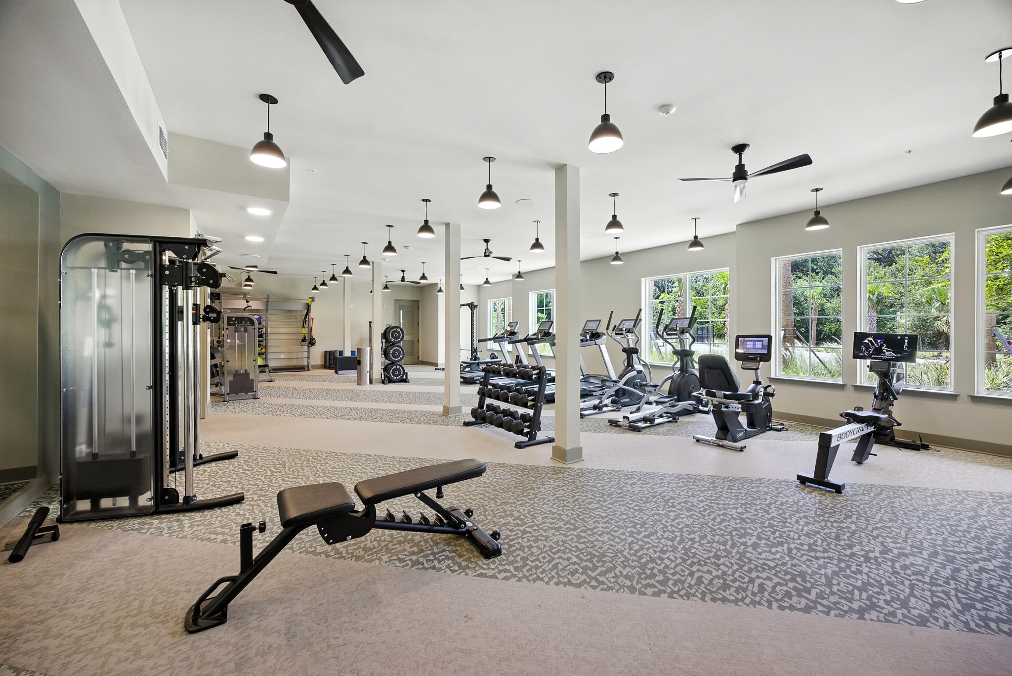 Modern gym with various exercise equipment including treadmills, stationary bikes, weight machines, and benches, in a spacious, well-lit room with large windows.