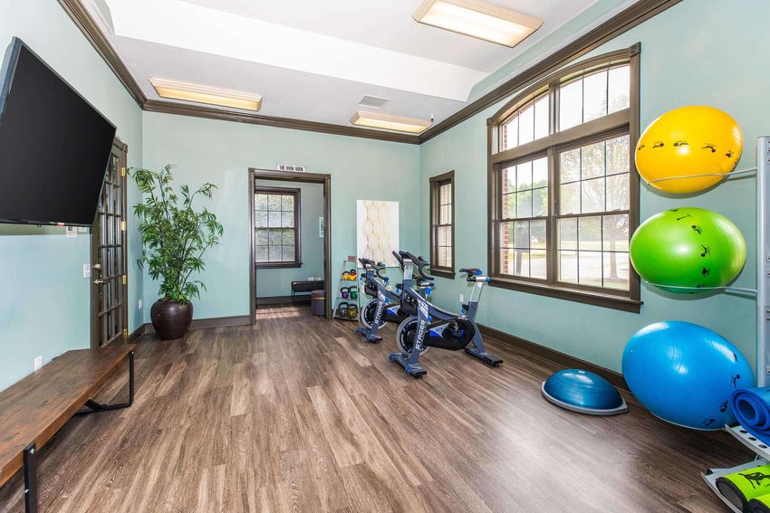 A fitness room featuring exercise equipment including stationary bikes, stability balls in blue and green, and a balance trainer. The room has large windows allowing natural light and a potted plant in the corner.