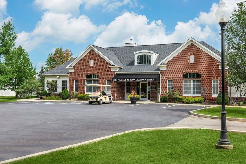 A brick building labeled "The Village of Oak Learning Center" with a well-maintained lawn, surrounded by trees and shrubs. A golf cart is parked in front, and there are light poles along the driveway under a partly cloudy sky.