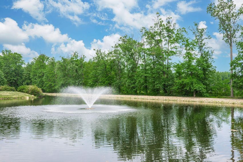 A serene pond surrounded by lush green trees, featuring a fountain spraying water in the center, with fluffy clouds in a blue sky reflected in the calm water.
