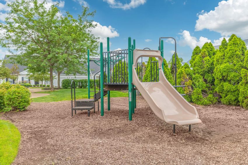 A playground featuring a beige slide and a small set of stairs, surrounded by green foliage and a blue sky with fluffy white clouds. The ground is covered in mulch, providing a safe play area.