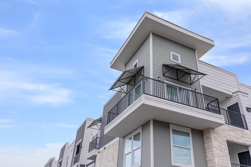 Modern apartment building with a sleek design, featuring multiple stories and balconies. The structure has a combination of light gray siding and stone accents, set against a clear blue sky.