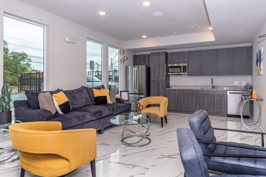 A modern living room featuring a gray sofa with yellow accent pillows, two yellow chairs, and a glass coffee table. The space has large windows allowing natural light, a sleek kitchen with gray cabinetry, and marble flooring, creating a stylish and inviting atmosphere.