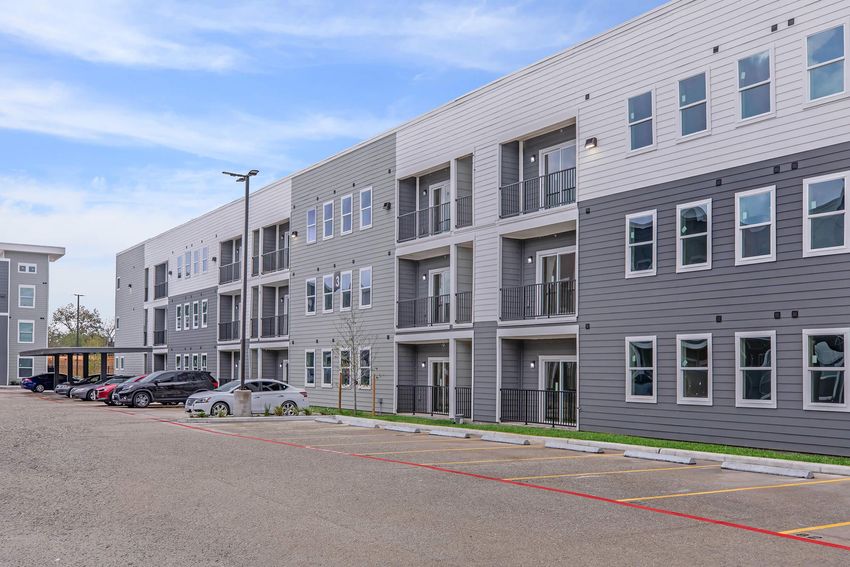 Modern apartment building featuring multiple stories with gray and white exterior. Balconies are visible on each unit, and there is a parking lot in front with several parked cars. The sky is clear, adding to the contemporary feel of the scene.