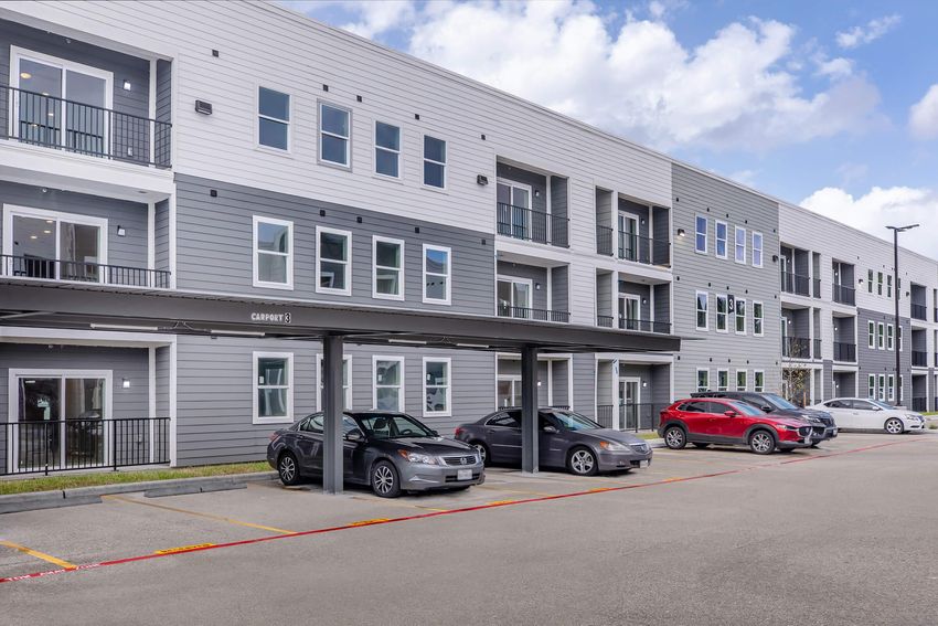 Exterior view of a modern apartment building featuring multiple gray and white facades, large windows, and a covered parking area with several parked cars. The sky is partly cloudy, contributing to a bright and inviting atmosphere.