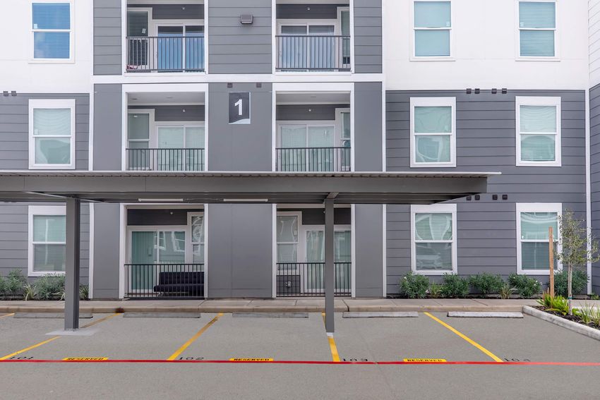 Exterior view of a modern apartment building with multiple levels. The facade features a mix of gray and white panels. Below, there is a covered parking area with marked spaces. The ground is paved, and small plants are visible beside the entrance. Windows are partially covered with curtains.