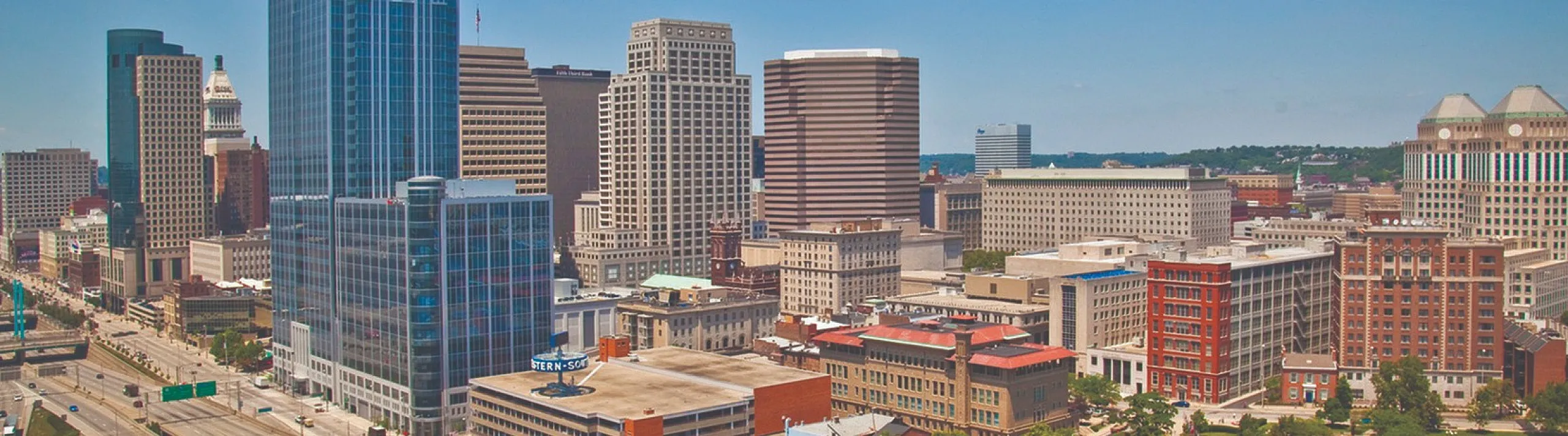 Vibrant city skyline with modern skyscrapers, lush green spaces, and a network of highways under a clear blue sky.
