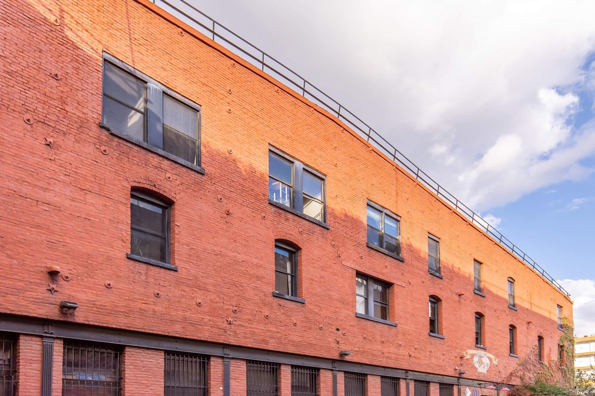 A brick building with multiple windows and a railing on the roof, set against a partly cloudy sky. The facade features a warm orange-red color and visible textures, with some windows reflecting light.