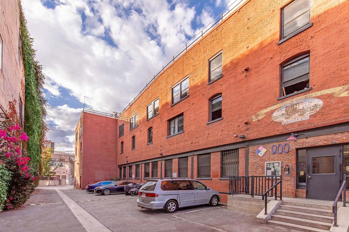 A narrow alleyway flanked by two brick buildings with large windows. One building features vibrant pink and green flowers climbing its wall. A gray minivan is parked in front, and the sky is partly cloudy. The scene has a rustic urban feel.