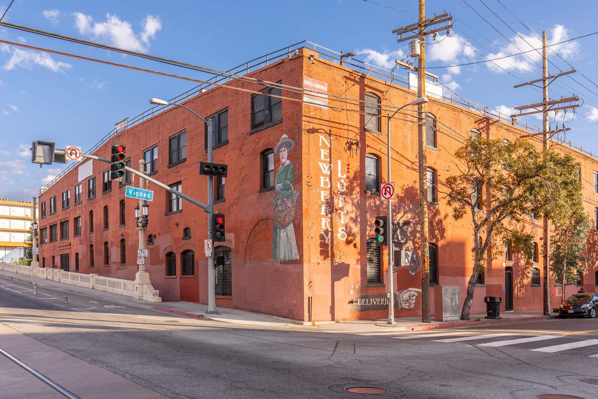 A red brick building on a street corner featuring a mural of a woman in a green dress. Traffic lights and street signs are visible. The sky is partly cloudy.