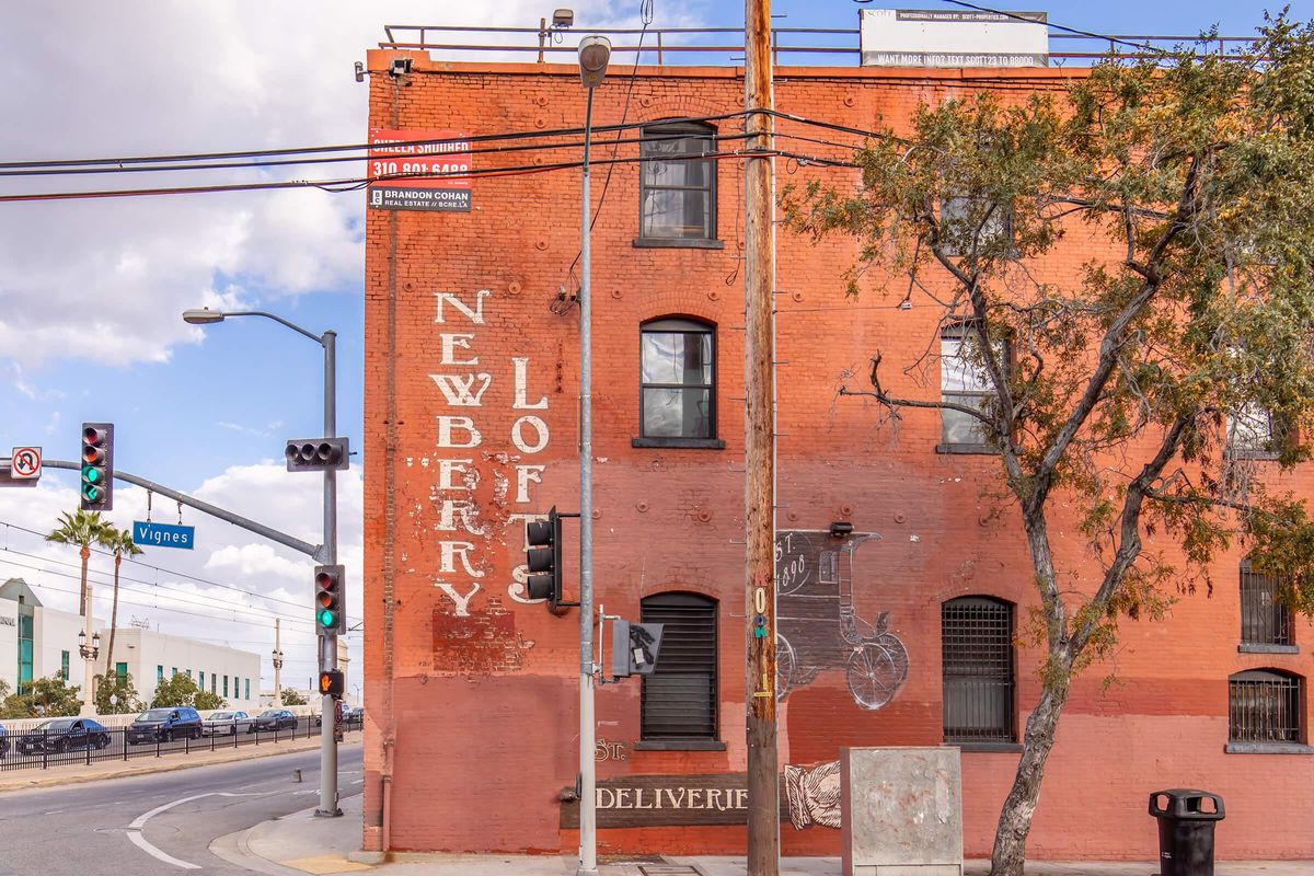 A brick building with the words "Newberry Loft" painted on the side. The building features large windows and has a vintage appearance. Traffic lights and power lines are visible in the foreground, along with a tree. The scene is set on a street with a partly cloudy sky.