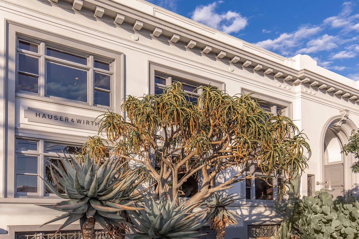 A modern building with large windows, featuring the name "HAUSER & WIRTH" above the entrance. In the foreground, there are various succulent plants and a large tree with thick branches and green leaves. The sky is partly cloudy, adding a blue hue to the scene.