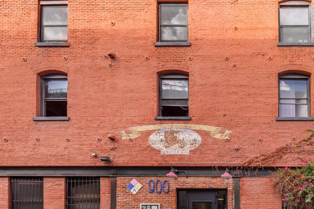 Red brick building facade featuring three windows on the upper level and a decorative mural with text on the lower portion. The building has black trim around the windows and a door, with hanging light fixtures and a small decorative element above the entrance.