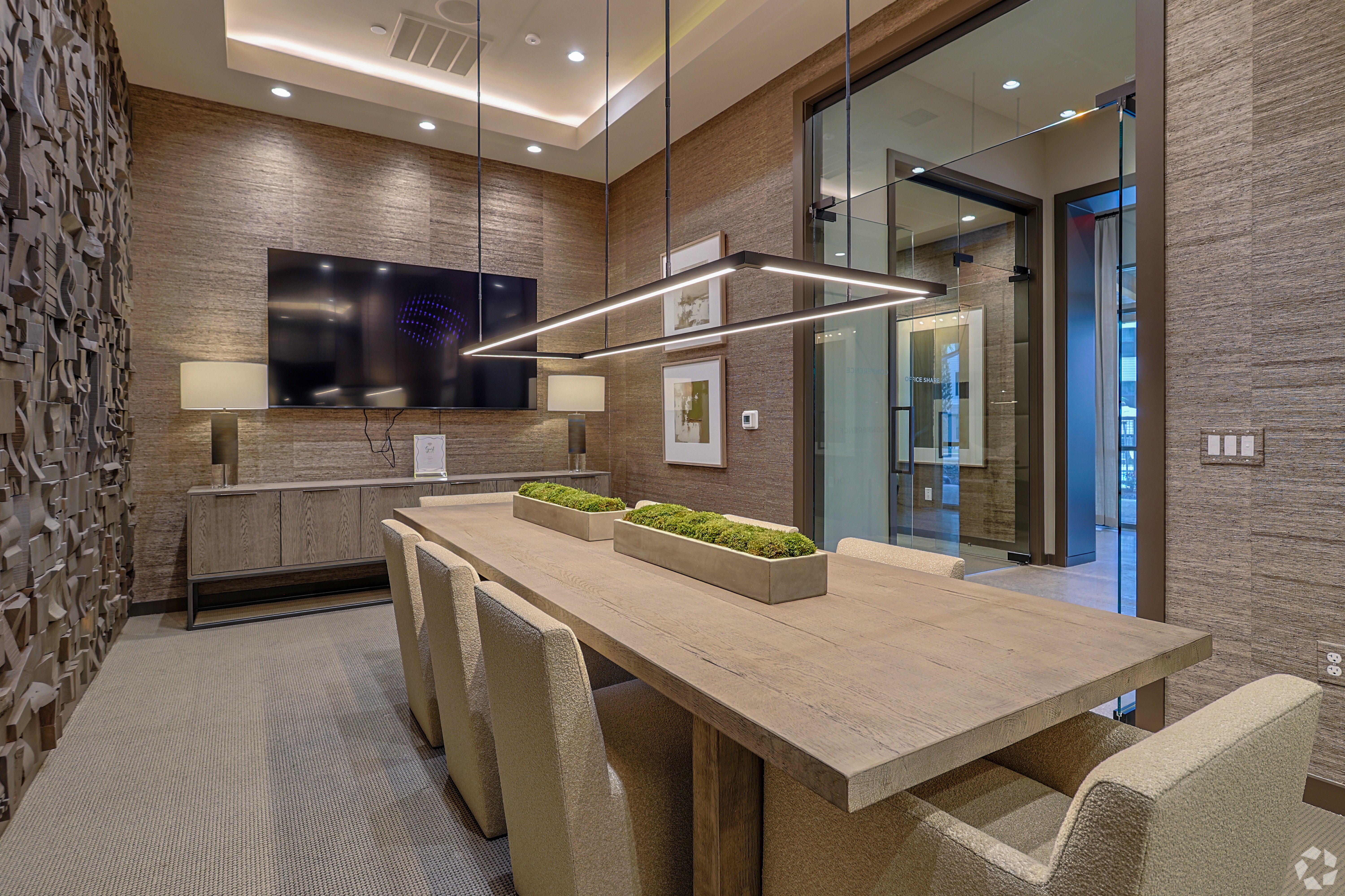 A modern conference room featuring a long wooden table with upholstered chairs, a flat-screen TV mounted on the wall, and decorative lighting. Greenery decorates the table, and there are textured wall panels in the background.