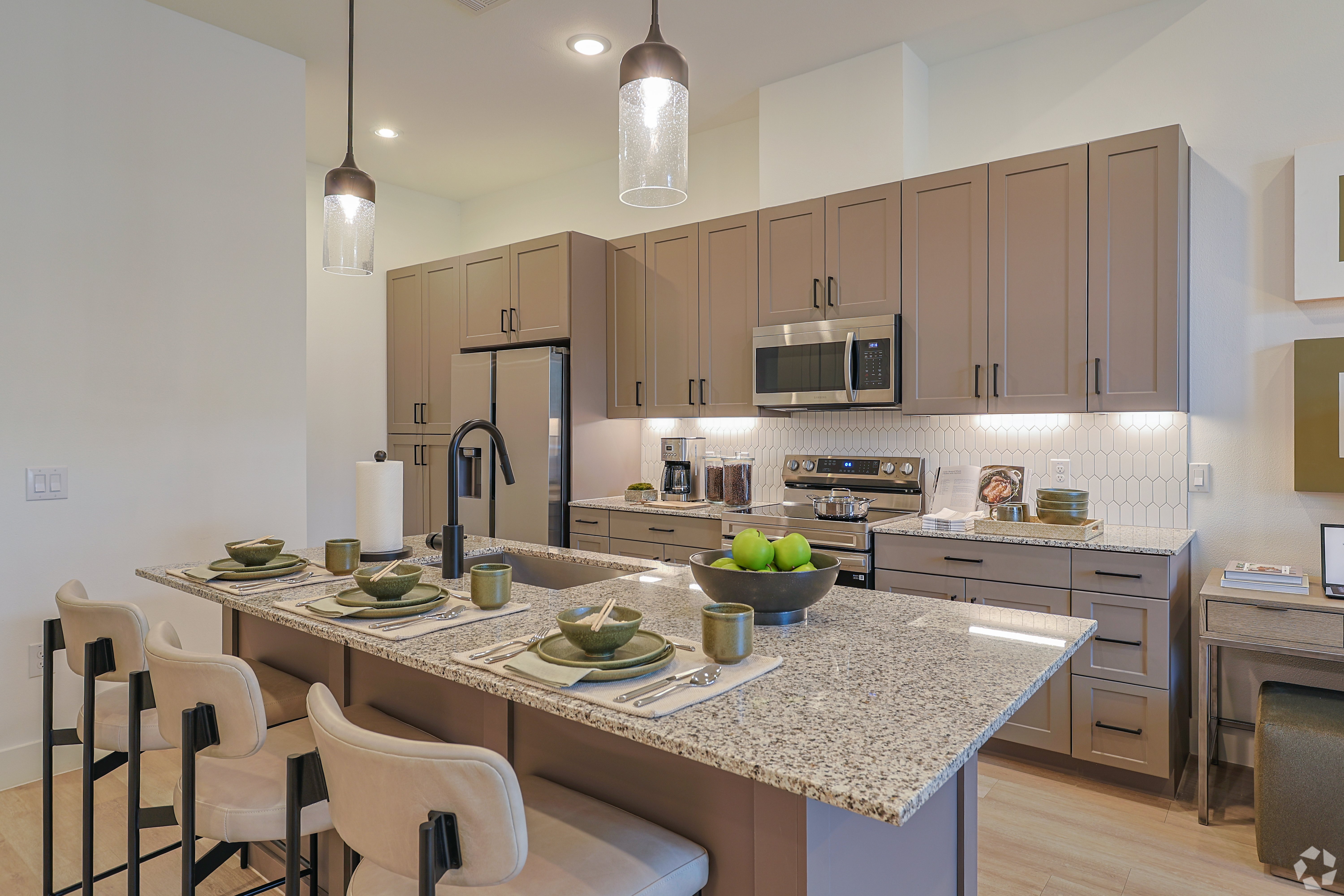A modern kitchen featuring brown cabinets, stainless steel appliances, and a large island with gray countertop. Four barstools are arranged at the island with plates and decorative items placed on the countertop. The kitchen is well-lit with pendant lights and the decor is minimalistic and stylish.