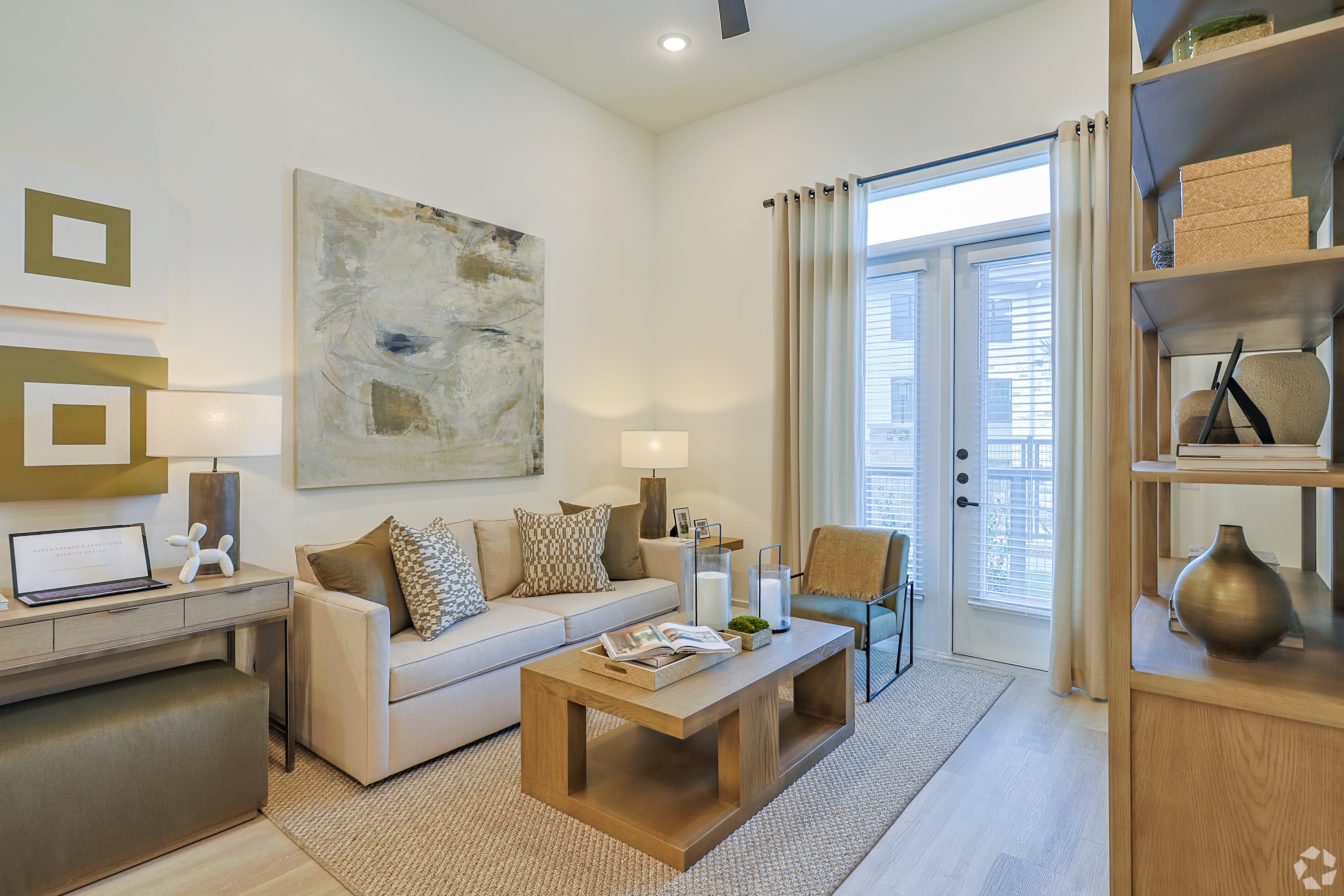 A modern living room featuring a light-colored sofa with decorative pillows, a wooden coffee table, and a minimalist desk. Nearby, there are large windows with sheer curtains letting in natural light, and abstract artwork on the wall. The decor includes earthy tones and light wood accents, creating a cozy atmosphere.